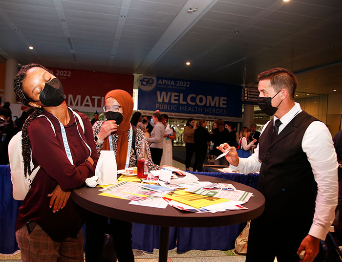 Two women wearing masks react in surprise when a magician reveals a card. 