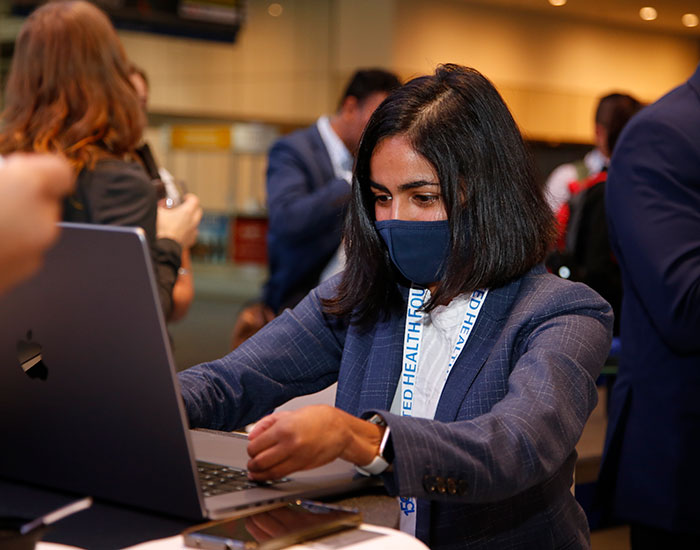 A woman in a blue blazer and white shirt and a black mask stands in front of a high-top table typing on her laptop. 