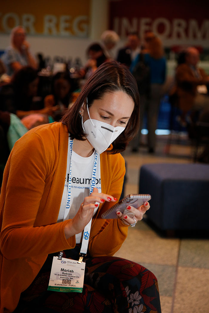 A woman in a dark orange cardigan and white mask sits on an ottoman looking down and typing on her smartphone. 