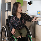 A South Asian person sits in her wheelchair and presses a button to lower the height-adjustable shelves in an accessible kitchen.