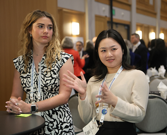 Young Asian woman is talking while moving her hands, while young white woman with hands clasped on hightop table looks at her. 