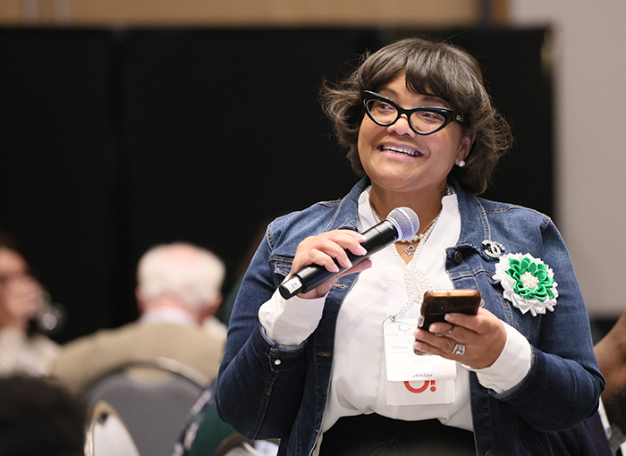 Woman in audience holds smartphone in left hand and microphone in right hand while smiling and asking a question.