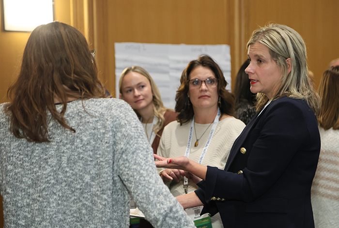 Woman in black blazer stands sideways at hightop table talking to three other women