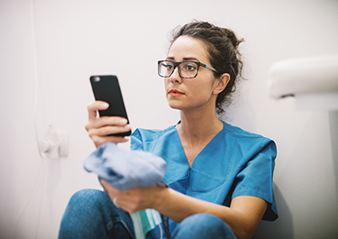 Person in scrubs sits on floor, leaning against the wall while reading their smartphone