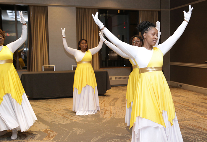 Four Black women in long white gloves and long white dresses with yellow overlays are doing a choreographed dance with their arms in the air. 