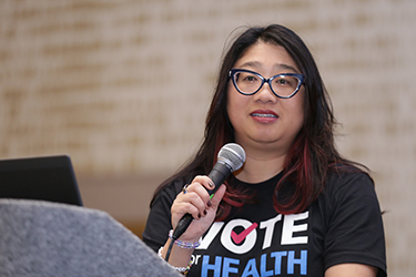 Past APHA President Chris Chanyasulkit wears a black T-shirt that says "Vote Health" while standing at a lectern and holding a microphone and talking to the crowd. 
