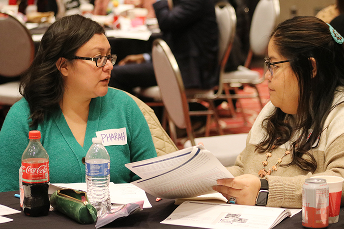 Two women sit together at a table with lots of paperwork and face each other while talking. 