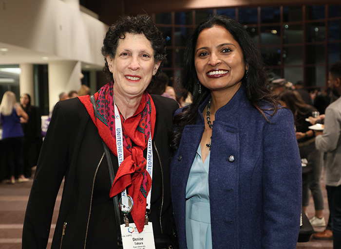 A woman in a black jacket with red neck scark and a woman in a blue jacket stand together and smile for the camera. 