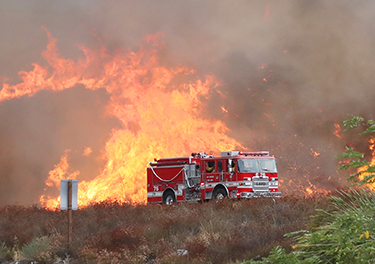 A fire truck is parked in front of extreme flames in an area of dried-out brush