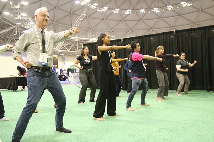 About 8 adults stand with knees bent, one arm pushed forward and one arm held back at their side, bent, during a taekwondo session in the Expo hall