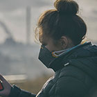 A woman wearing a mask and a winter coat sits outside checking air quality measures on her cellphone while smoke billows from a factory in the distance. 